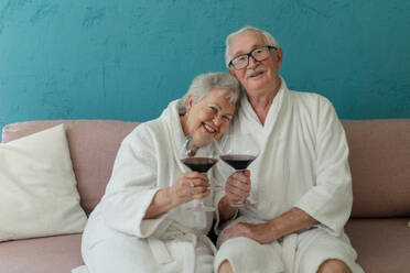 Glückliches älteres Paar, das zusammen im Bademantel auf dem Sofa mit einem Glas Wein sitzt und eine schöne Zeit zu Hause hat. - HPIF11425