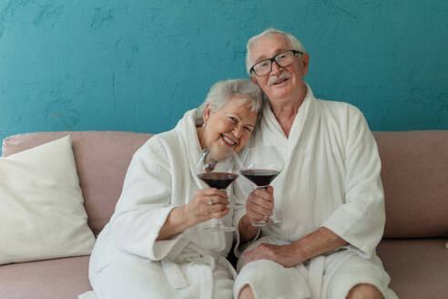 Glückliches älteres Paar, das zusammen im Bademantel auf dem Sofa mit einem Glas Wein sitzt und eine schöne Zeit zu Hause hat. - HPIF11425