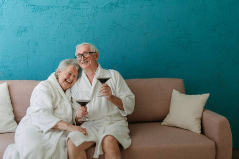 Glückliches älteres Paar, das zusammen im Bademantel auf dem Sofa mit einem Glas Wein sitzt und eine schöne Zeit zu Hause hat. - HPIF11426