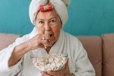 Ein glückliches älteres Ehepaar sitzt im Bademantel auf einem Sofa und sieht mit Popcorn fern. - HPIF11441