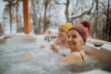 Älteres Paar mit Zipfelmütze, das an einem kalten Wintertag auf der Terrasse ein Glas Wein genießt und sich in der Badewanne erfrischt. - HPIF11490