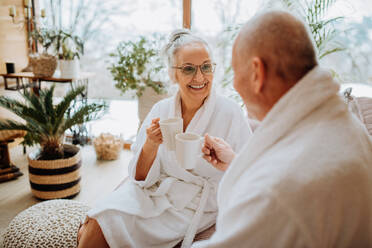 Ein älteres Paar in Bademänteln genießt die gemeinsame Zeit in ihrem Wohnzimmer, trinkt einen heißen Tee und genießt die ruhige und gemütliche Atmosphäre. - HPIF11517