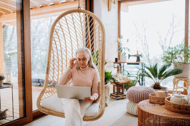 Ältere Frau, die in einer Schaukel sitzt und an einem Laptop arbeitet, Konzept des Home Office und des aktiven Lebensstils älterer Menschen. - HPIF11519