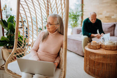 Ältere Frau sitzt in einer Schaukel im Freien und arbeitet am Laptop, ihr Mann ruht sich auf einem Sofa aus. Konzept des Home Office und des aktiven Lebensstils älterer Menschen. - HPIF11521