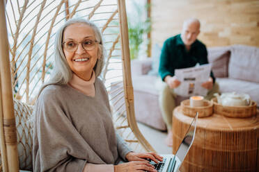 Ältere Frau sitzt in einer Schaukel im Freien und arbeitet am Laptop, ihr Mann ruht sich auf einem Sofa aus. Konzept des Home Office und des aktiven Lebensstils älterer Menschen. - HPIF11523