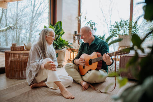 Älterer Mann spielt auf der Gitarre seiner Frau, die in einem gemütlichen Wohnzimmer sitzt und den Herbsttag genießt. - HPIF11528