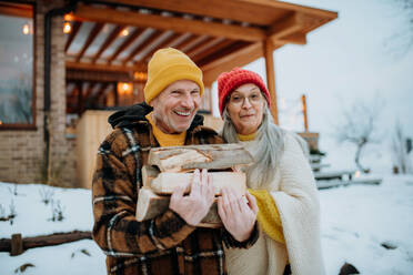 Ein älteres Ehepaar hält an einem Wintertag Holzscheite vor seinem Haus. - HPIF11544