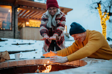 Älteres Ehepaar, das an einem Winterabend gemeinsam an einer Feuerstelle im Freien sitzt und heizt. - HPIF11554