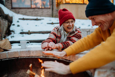 Älteres Ehepaar, das an einem Winterabend gemeinsam an einer Feuerstelle im Freien sitzt und heizt. - HPIF11555