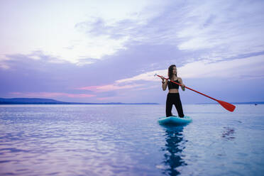 A young beautiful girl surfer paddling on surfboard on the lake at sunrise - HPIF11590