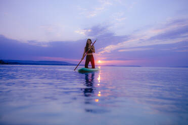 A young beautiful girl surfer paddling on surfboard on the lake at sunrise - HPIF11598