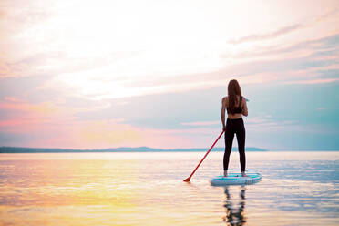 Rückansicht einer Surferin, die auf einem Surfbrett auf dem See bei Sonnenaufgang paddelt. - HPIF11602