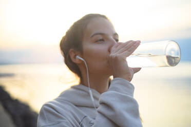A young beautiful sportive girl listening to music and drinking water at sunrise by the lake. - HPIF11605