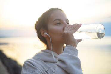 Ein junges, schönes, sportliches Mädchen, das bei Sonnenaufgang am See Musik hört und Wasser trinkt. - HPIF11605