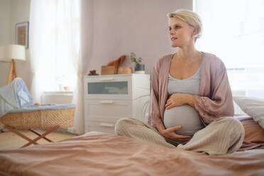 Glückliche schwangere Frau, die auf einem Bett sitzend ihren Bauch streichelt. - HPIF11610