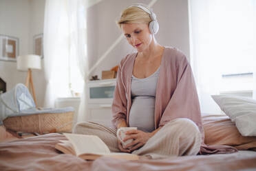 Schwangere Frau, die in einem Bett sitzt, Musik hört und ein Buch liest und die Zeit für sich genießt. - HPIF11611