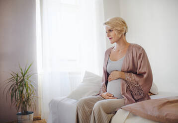 Glückliche schwangere Frau, die auf einem Bett sitzend ihren Bauch streichelt. - HPIF11637