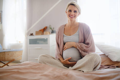 Glückliche schwangere Frau, die auf einem Bett sitzend ihren Bauch streichelt. - HPIF11640