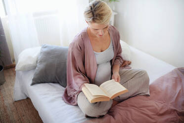 Schwangere Frau sitzt auf dem Bett und liest ein Buch. - HPIF11642