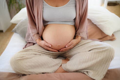 Glückliche schwangere Frau, die auf einem Bett sitzend ihren Bauch streichelt. - HPIF11643