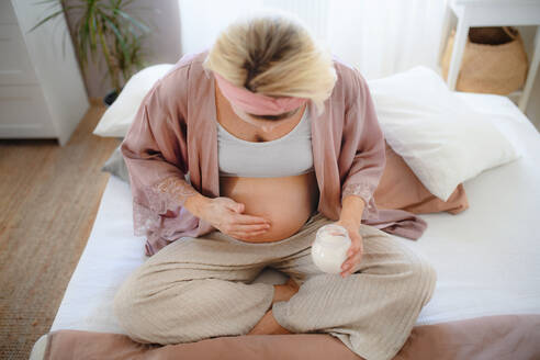 Schwangere Frau, die ihren Bauch eincremt, in einem Bett sitzend, sich um sich selbst kümmernd. - HPIF11645