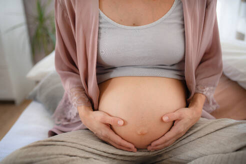 Glückliche schwangere Frau, die auf einem Bett sitzend ihren Bauch streichelt. - HPIF11646