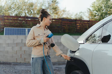 Junge Frau hält das Stromversorgungskabel ihres Autos in der Hand, um es zu Hause aufzuladen, nachhaltiges und wirtschaftliches Verkehrskonzept. - HPIF11700