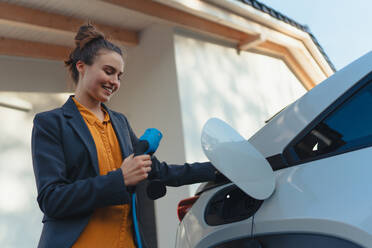 Junge Frau hält das Stromversorgungskabel ihres Autos in der Hand, um es zu Hause aufzuladen, nachhaltiges und wirtschaftliches Verkehrskonzept. - HPIF11708