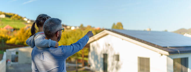 Rückansicht des Vaters, der seine kleine Tochter in den Armen hält und ihr Haus mit Sonnenkollektoren zeigt: Alternative Energie, Ressourceneinsparung und nachhaltiger Lebensstil. - HPIF11773