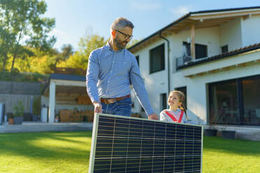 Vater mit seiner kleinen Tochter, die ein Solarmodul im Hinterhof trägt. Alternative Energie, Ressourcenschonung und nachhaltiger Lebensstil. - HPIF11793