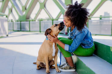 Multirassisches Mädchen sitzt und ruht sich mit ihrem Hund draußen auf der Brücke aus, trainiert ihn, verbringt die Freizeit zusammen. Konzept der Beziehung zwischen einem Hund und Teenager, Alltag mit Haustier. - HPIF11859