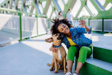 Multiracial girl sitting and resting with her dog outside in the bridge, taking selfie with him, spending leisure time together. Concept of everyday life with pet. - HPIF11866