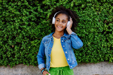 Multirassisches Mädchen genießt Musik in den Kopfhörern, stehend vor einem grünen Busch, Porträt. - HPIF11873