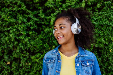 Multirassisches Mädchen genießt Musik in den Kopfhörern, stehend vor einem grünen Busch, Porträt. - HPIF11874