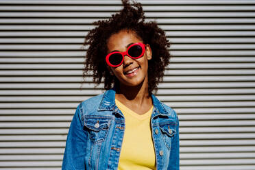 Porträt eines jungen glücklichen multirassischen Mädchens mit roter Sonnenbrille und Afrofrisur, stehend im Freien. - HPIF11877