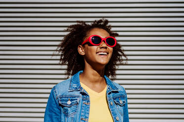 Porträt eines jungen glücklichen multirassischen Mädchens mit roter Sonnenbrille und Afrofrisur, stehend im Freien. - HPIF11878