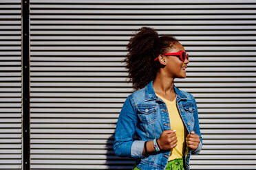 Porträt eines jungen glücklichen multirassischen Mädchens mit roter Sonnenbrille und Afrofrisur, stehend im Freien. - HPIF11880