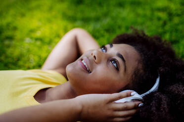 Multirassisches Mädchen liegt im Gras und genießt Musik aus Kopfhörern, Seitenansicht. - HPIF11890