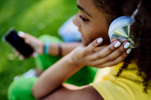 Multirassisches Mädchen, das im Gras sitzt und Musik mit Kopfhörern genießt, hoher Blickwinkel. - HPIF11896