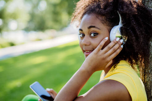Multirassisches Mädchen sitzt im Gras und genießt Musik mit Kopfhörern, Seitenansicht. - HPIF11897