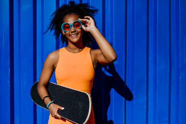 Multirassisches Teenager-Mädchen mit Skateboard vor einer blauen Wand an einem Sommertag. - HPIF11898