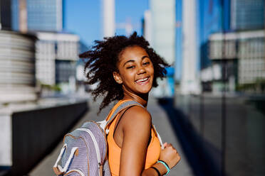Multiracial teenage girl walking with a backpack in modern city centre during summer day, back to school concept. - HPIF11900