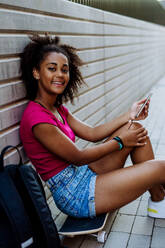 Multiracial teenage girl with backpack sitting at skateboard, using phone, during a summer day. - HPIF11918