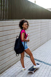 Multirassisches Teenager-Mädchen mit Rucksack am Skateboard, an einem Sommertag. - HPIF11919