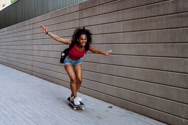 Multirassisches Teenager-Mädchen fährt Skateboard vor einer Betonmauer, balancierend, Seitenansicht. - HPIF11920