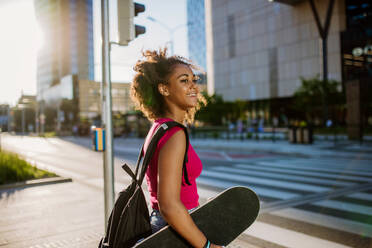 Multiracial Teenager-Mädchen mit Rucksack und Skateboard, zu Fuß in der Stadt während eines Sommertages. - HPIF11921