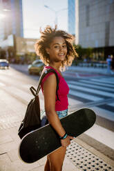 Multiracial teenage girl with backpack and skateboard, walking in city during a summer day. - HPIF11922