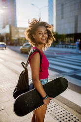 Multiracial Teenager-Mädchen mit Rucksack und Skateboard, zu Fuß in der Stadt während eines Sommertages. - HPIF11922