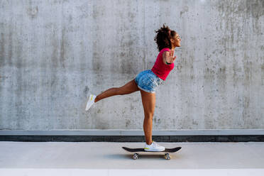 Multirassisches Teenager-Mädchen fährt Skateboard vor einer Betonmauer, steht auf einem Bein und balanciert, Seitenansicht. - HPIF11924