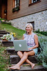 Eine ältere Frau benutzt einen Laptop und bearbeitet Bestellungen für ihre selbst angebauten Bio-Blumen und Gemüse im Garten - HPIF11979